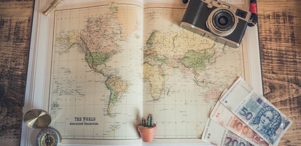atlas showing old maps of the world with camera, euros and compass sitting on top of atlas on wooden table